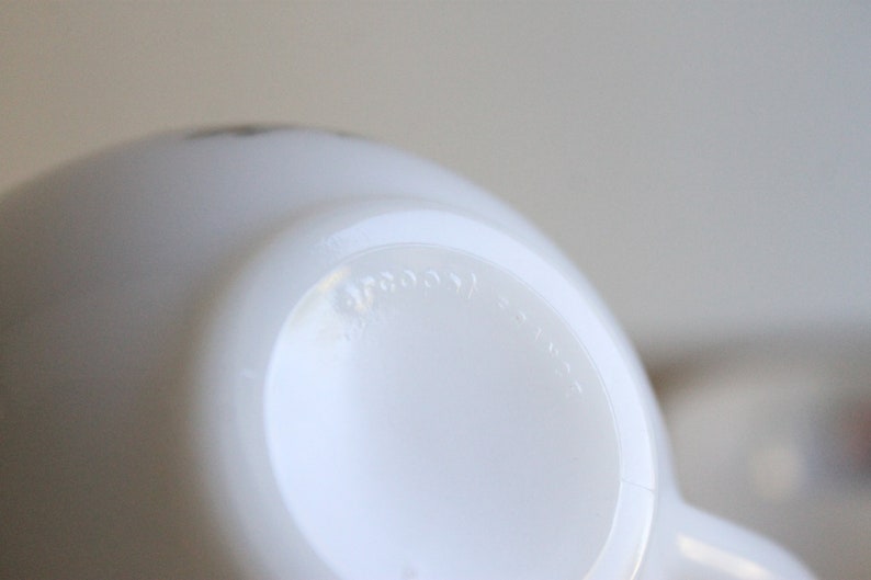 May include: Close-up of a white, opaque, ceramic mug. The bottom of the mug is visible, with the word "arcoos" embossed on the base. The mug has a rounded shape and a handle is partially visible.