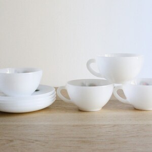 May include: A set of white ceramic teacups and saucers. The cups have a rounded shape with a handle, and the saucers are flat. The teacups and saucers are arranged on a light-colored wooden surface.