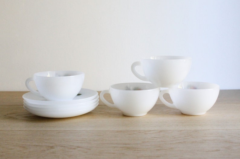 May include: A set of white ceramic teacups and saucers. The cups have a rounded shape with a handle, and the saucers are flat. The teacups and saucers are arranged on a light-colored wooden surface.