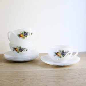 May include: Set of four white ceramic teacups with a floral design featuring pears and grapes. The cups are stacked on a saucer.