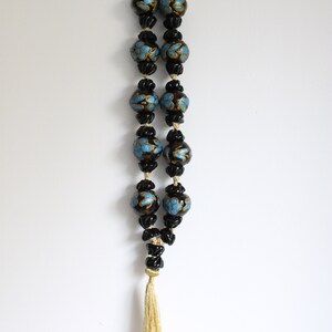 Puede incluir: Un collar de cuentas negras con detalles azules y dorados y un borla dorada. El collar est&aacute; colgado de un gancho en una pared blanca.