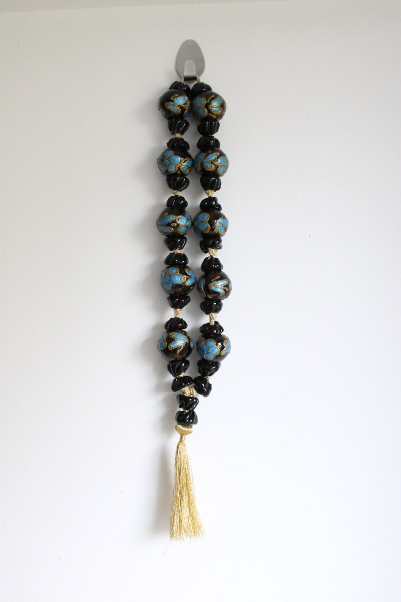 Puede incluir: Un collar de cuentas negras con detalles azules y dorados y un borla dorada. El collar est&aacute; colgado de un gancho en una pared blanca.
