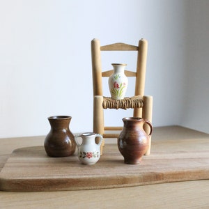 May include: Four small ceramic vases, two brown and two white with floral designs, on a wooden chopping board. One white vase sits on a small wooden chair.