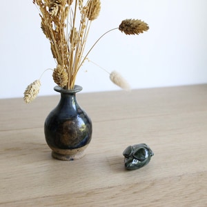 Peut inclure: Un petit vase en céramique bleu foncé avec un glaçage brun posé sur une surface en bois. Le vase est rempli d'herbes séchées et d'une seule fleur séchée. Une petite figurine de souris en céramique verte est posée sur la surface à côté du vase.
