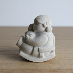 Op de afbeelding: Een lichtgekleurde, steenachtige sculptuur van een persoon die een kleiner figuur vasthoudt. De figuren zitten op een schommelbasis. De sculptuur heeft een matte afwerking en een neutrale achtergrond.