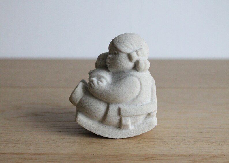 Op de afbeelding: Een lichtgekleurde, steenachtige sculptuur van een persoon die een kleiner figuur vasthoudt. De figuren zitten op een schommelbasis. De sculptuur heeft een matte afwerking en een neutrale achtergrond.