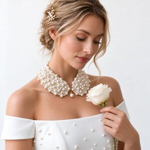 May include: A detachable collar made of clusters of white pearls, featuring a single dangling pearl at the center. The collar is worn over a white off-the-shoulder dress embellished with scattered white pearls and a matching pearl hair accessory.