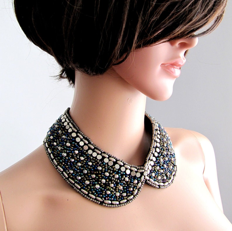 May include: A silver and blue beaded collar necklace with a wide, rounded shape. The necklace is made of small, round beads and larger, flat, silver beads.