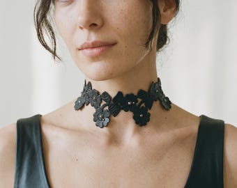 May include: A black choker necklace featuring a series of interconnected floral and leaf shapes. The design includes small metallic studs at the centre of each flower, creating a detailed botanical pattern that rests closely around the neck.
