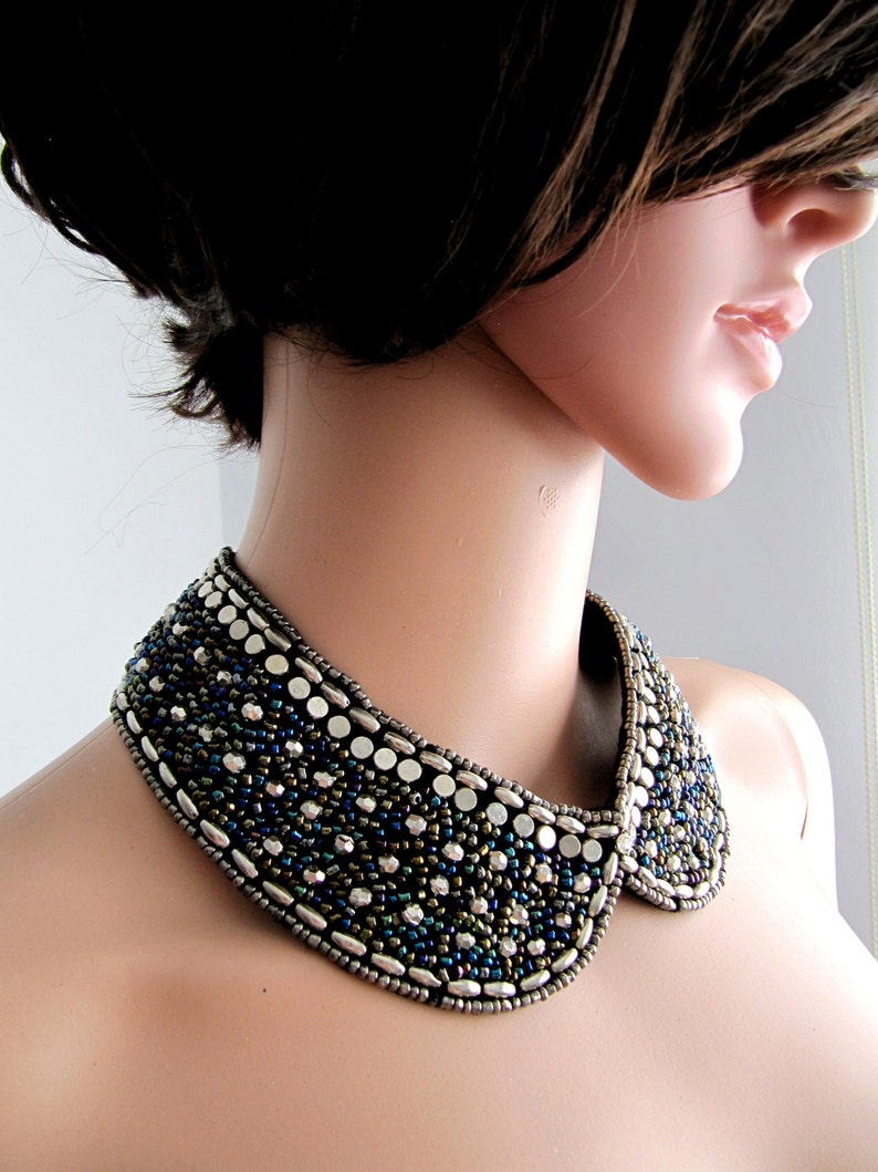 May include: A beaded collar necklace with a wide, rounded shape. The necklace is made of silver, blue, and green beads.