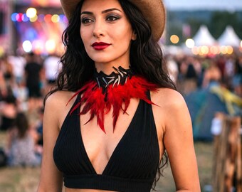 May include: A tan cowboy hat worn with a black ribbed halter crop top and a choker necklace featuring black lace, silver-toned spikes, and vibrant red feathers. The outfit is paired with blue denim jeans, creating a distinct look.