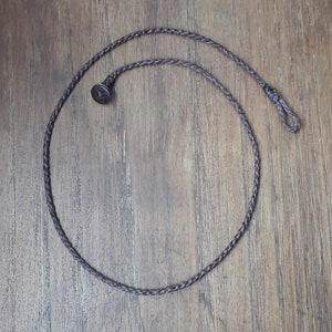 Puede incluir: Un collar de cordón trenzado marrón con un cierre de metal. El collar está sobre una superficie de madera.