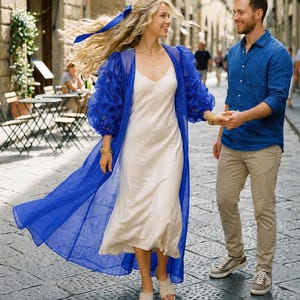 May include: A cream-colored slip dress paired with a sheer, long-sleeved royal blue organza duster coat. The ensemble is worn with beige slide sandals and gold bracelets. The blue coat features voluminous sleeves and a flowing, lightweight fabric.