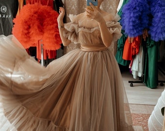 May include: A floor-length beige tulle dress featuring an off-the-shoulder neckline with ruffled sleeves, a fitted satin waistband, and a voluminous skirt with a wide ruffled hemline. The dress is constructed from multiple layers of sheer fabric.