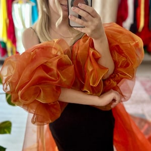 Può includere: Una donna indossa un abito nero con un lungo strato di tulle arancione e oro. Il tulle ha maniche grandi e arricciate.
