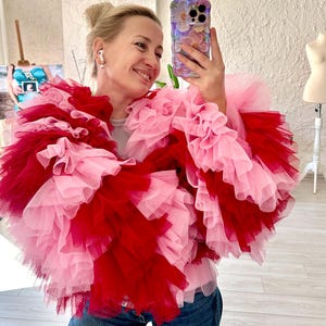 May include: A woman wearing a layered, ruffled garment in shades of pink and red. The garment appears to be made of tulle or a similar fabric, creating a voluminous, textured effect. The woman is smiling and holding a phone.