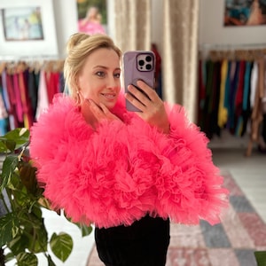 May include: A woman wearing a bright pink tulle top with a black skirt. She is taking a selfie with a smartphone.
