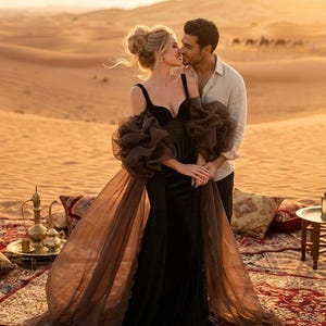 May include: A couple embraces in a desert setting. The woman wears a black velvet gown with sheer brown sleeves and a flowing train. The man wears a light-colored shirt. The scene is set on a patterned rug with pillows and Middle Eastern decor.