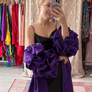 May include: A woman in a black dress and purple overcoat with large, ruffled sleeves poses in a boutique. The overcoat has a long train. A variety of colorful dresses hang on racks in the background. A gold-framed mirror is on the right.