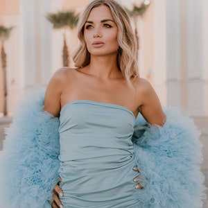 May include: A woman wearing a light blue strapless dress with a matching light blue tulle shrug. The dress is fitted and has a ruched design. The shrug is fluffy and has a lot of volume.