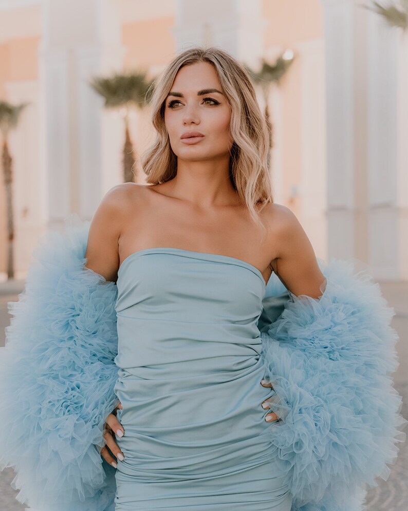 May include: A woman wearing a light blue strapless dress with a matching light blue tulle shrug. The dress is fitted and has a ruched design. The shrug is fluffy and has a lot of volume.