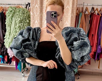 May include: A woman wearing a black dress with a blue and silver jacket featuring large, puffy sleeves. The jacket has a textured, almost quilted appearance. The woman is holding a smartphone in front of her face.