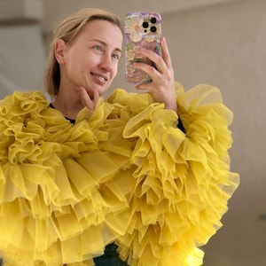 May include: A bright yellow, ruffled garment with multiple layers of fabric. The garment is worn over a dark top. The person is holding a phone with a floral phone case.