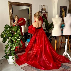 May include: A floor-length red gown featuring voluminous puff sleeves and a strapless bodice. The garment is crafted from a sheer, lightweight fabric that creates a dramatic train, flowing outward from the waist to the floor in a wide, full silhouette.