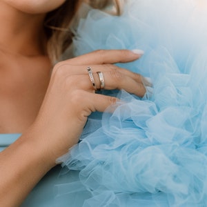 May include: A woman's hand wearing two thin gold rings with small diamonds. The hand is resting on a light blue tulle fabric.