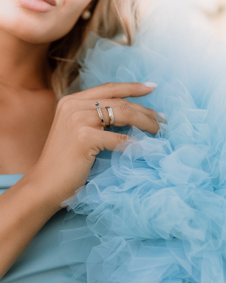 May include: A woman's hand wearing two thin gold rings with small diamonds. The hand is resting on a light blue tulle fabric.