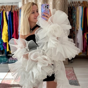 May include: A woman wearing a black dress and a large white ruffled garment. The garment has multiple layers of flowing fabric. The woman is holding a phone in front of her face, and the background shows clothing racks.