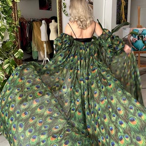 May include: A long, flowing dress with a peacock feather design in green, blue, and gold. The dress has off-the-shoulder sleeves and a black bodice. The model is viewed from the back, highlighting the dress's dramatic, floor-length design.
