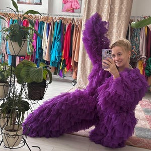 May include: A woman poses in a full-length, ruffled purple gown. The gown is made of layers of fabric, creating a voluminous effect. The background includes racks of colorful clothing and potted plants.