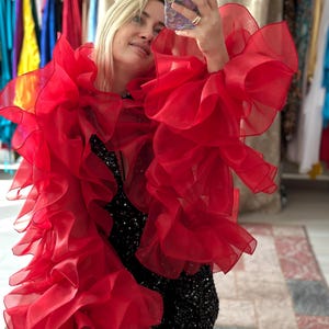 Puede incluir: Una llamativa blusa de organza roja con volantes y mangas voluminosas. Se lleva sobre un vestido negro con lentejuelas. El fondo muestra percheros, lo que sugiere un entorno de moda o venta al por menor.
