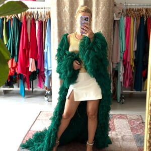 May include: A woman wearing a long, emerald green, ruffled robe over a yellow top and white shorts. The robe has a train and is made of a sheer fabric. She is wearing jeweled sandals. Clothing items hang on racks in the background.