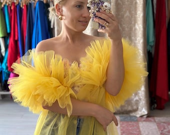 May include: A woman wearing a bright yellow tulle dress with a ruffled neckline and sleeves. The dress is paired with blue jeans. She is holding a smartphone with a decorative case. Gold earrings are visible.