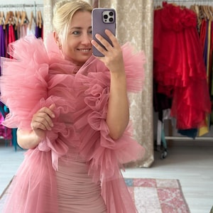 Pode incluir: Uma mulher vestindo um vestido de tule rosa e um xale com babados combinando. O vestido é rosa claro e tem um design franzido. O xale é volumoso e adiciona um toque dramático ao look. O fundo mostra araras de roupas e um tapete.