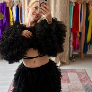 May include: Black ruffled crop top and skirt set. The top features voluminous, layered ruffles on the sleeves and bodice. The skirt has a tiered, ruffled design. The outfit is worn by a person taking a selfie in a room with clothing racks.