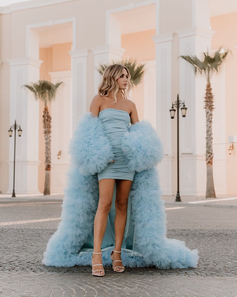 May include: A woman wearing a light blue strapless dress with a matching tulle coat. The coat has a long train and is very fluffy. She is standing on a paved surface in front of a white building.