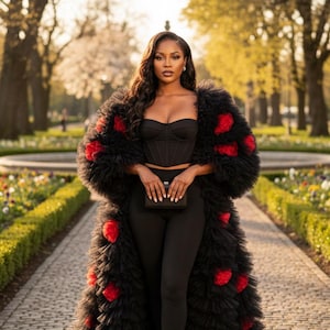 May include: A woman in a black outfit, including a corset top and leggings, is wearing a long, black coat adorned with red accents. She is holding a small black clutch and wearing black heels. The background features a paved walkway and a park setting.