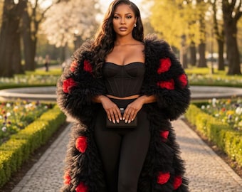 May include: A woman in a black outfit, including a corset top and leggings, is wearing a long, black coat adorned with red accents. She is holding a small black clutch and wearing black heels. The background features a paved walkway and a park setting.