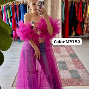 Puede incluir: Un vibrante conjunto de tul magenta, con un top con volantes adornado con adornos florales y una capa larga a juego. El atuendo se combina con vaqueros de mezclilla desgastados. El texto "Color MY182" es visible.