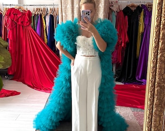 May include: A woman in a mirror wearing a white top and pants, and a long, turquoise tulle coat. The background shows a clothing store with racks of colorful dresses and gowns. A gold-framed mirror reflects the scene.