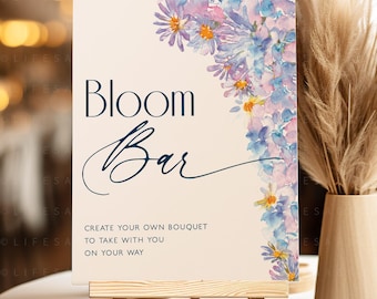 May include: A sign with the text "Bloom Bar" in blue and white lettering, with a watercolor floral design in shades of purple and blue. The text below reads "Create your own bouquet to take with you on your way."