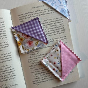 May include: A collection of handmade fabric corner bookmarks in various patterns, including floral, gingham, and polka dots. The bookmarks are placed on the pages of an open book, showcasing their decorative and functional design.