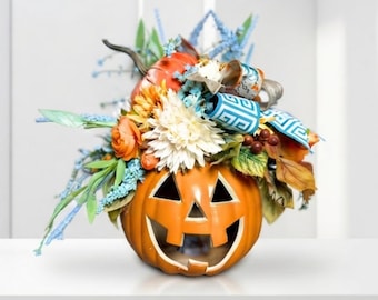 Puede incluir: Una calabaza de cerámica con cara tallada, decorada con un arreglo de flores artificiales naranjas y blancas, follaje verde, bayas marrones y una cinta azul y blanca con un diseño geométrico de greca griega.
