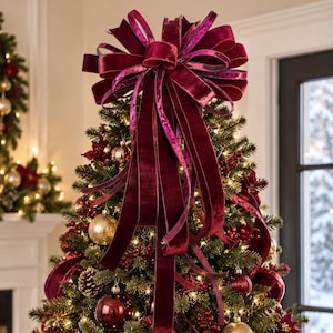 May include: A large, deep burgundy velvet bow tree topper with multiple cascading loops and long, flowing ribbon tails. The fabric has a soft, plush texture that catches the light, creating subtle highlights across the dark red surface.