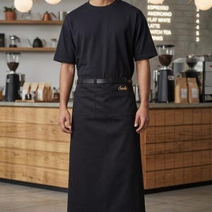 Puede incluir: Una persona con una camiseta negra y un delantal largo negro con el nombre "Sade" bordado en dorado. El delantal tiene dos bolsillos delanteros y un cinturón negro. El fondo es una cafetería.