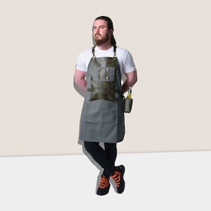 May include: A gray apron with a dark green leather chest pocket and accents. The apron has two large front pockets and a small pocket for a beverage. The apron is designed for grilling or cooking.