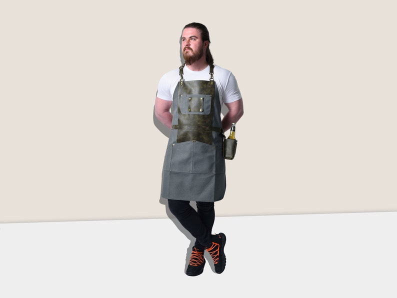 May include: A gray apron with a dark green leather chest pocket and accents. The apron has two large front pockets and a small pocket for a beverage. The apron is designed for grilling or cooking.